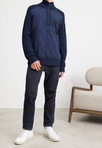 Pull-over matelassé bleu marine avec col montant, demi-fermeture éclair, manches côtelées, associé à un jean foncé et des baskets blanches. Design simple et minimaliste.
