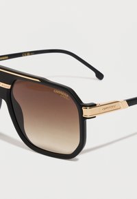 Occhiali da sole neri con finitura opaca, accenti dorati sulle tempie, lenti marroni a gradiente e il nome del marchio "CARRERA" inciso sulla montatura.