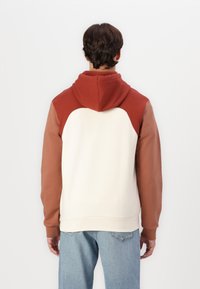 Felpa con cappuccio dal corpo crema, spalle e cappuccio rosso ruggine, e maniche lunghe color marrone chiaro. Tessuto morbido con polsini e orlo a coste.