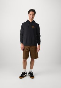 Zwarte hoodie met een voorzak en kleurrafiek, gecombineerd met kaki shorts en zwarte sneakers met witte accenten en gumzolen.