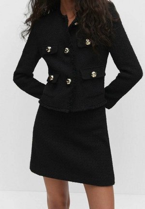 Femme portant une veste noire texturée avec des boutons dorés et une jupe noire assortie sur un fond uni.