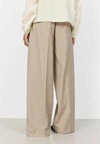 Beige weit geschnittene Hose aus einem glatten Stoff, mit einem hoch sitzenden Design und einer lockeren Passform. Der Saum ist leicht zulaufend.