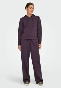 Fioletowa bluza z kapturem na zamek błyskawiczny, z sznurkiem i dopasowanymi, luźnymi spodniami. Miękka tekstura, ściągacze na mankietach i kieszenie boczne. Sneakersy dopełniają stylizację.