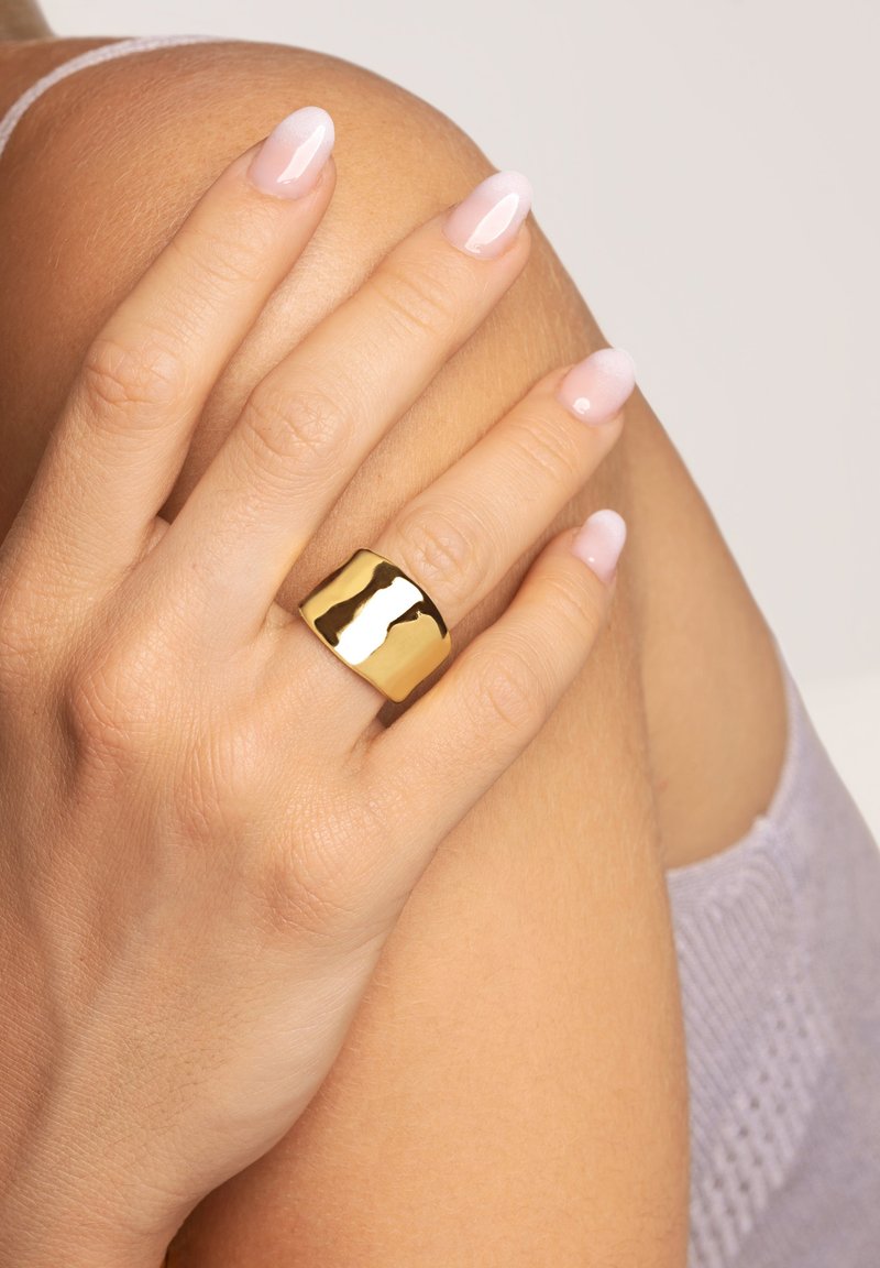 Anillo grueso de tono dorado con una superficie lisa y reflectante, que se lleva en una mano apoyada sobre un hombro, mostrando la textura natural de la piel.