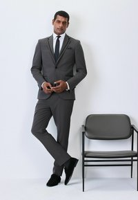 Traje gris a medida con un diseño ajustado, camisa blanca, corbata negra y zapatos de ante negros. Apoyado contra una silla negra minimalista.