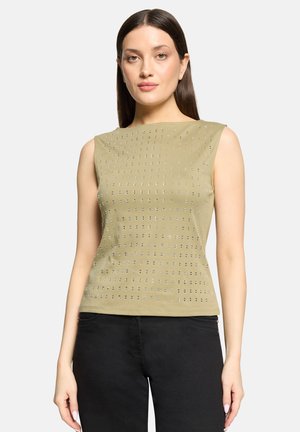 Vrouw met recht bruin haar draagt een mouwloze olijfgroene top met reflecterende vierkante patronen en zwarte broek, staand tegen een effen achtergrond.