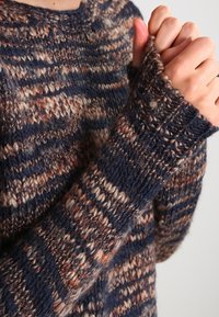 Sweter dzianinowy z mieszanką ciemnoniebieskich, brązowych i kremowych pasków. Gruba włóczka nadaje mu teksturowany wygląd z ściągaczami przy mankietach.