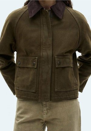 Veste marron avec col marron foncé et poches avant à rabat portée sur un pantalon beige, personne debout avec les mains derrière le dos.