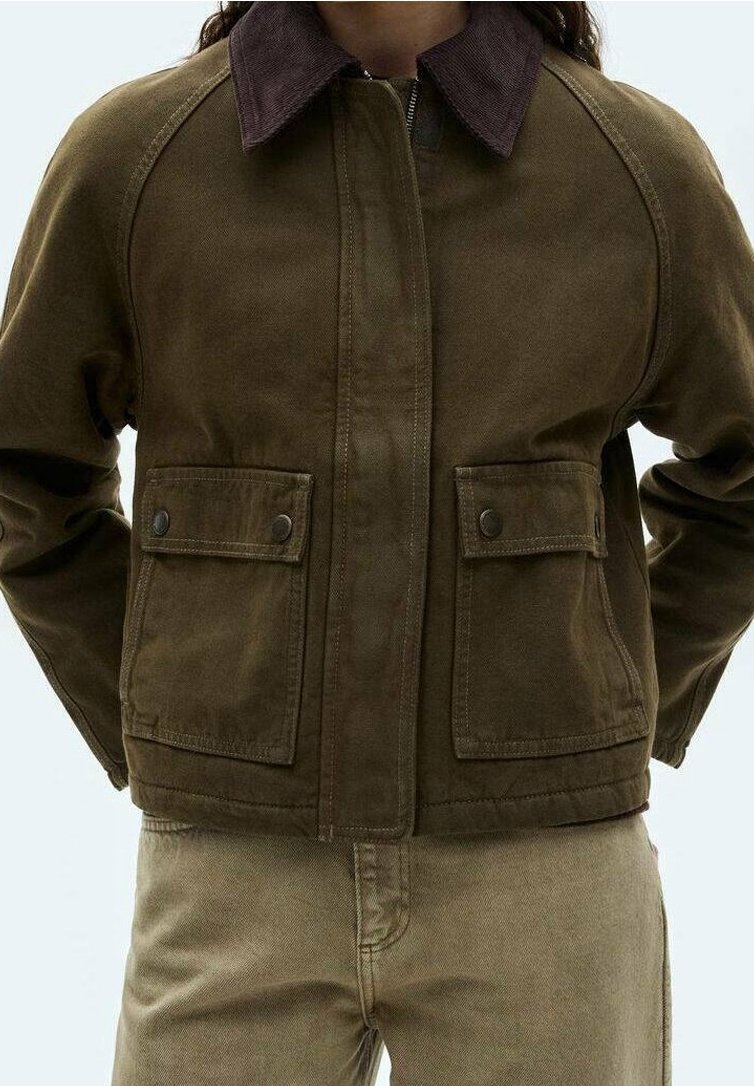 Veste marron avec col marron foncé et poches avant à rabat portée sur un pantalon beige, personne debout avec les mains derrière le dos.
