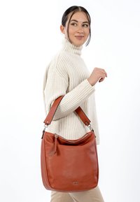 Braune Leder-Umhängetasche mit einem Reißverschluss oben, glatter Oberfläche und breitem verstellbarem Riemen, gehalten von einer Person mit einem cremefarbenen Pullover.