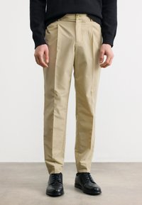 Άνδρας που φοράει μπεζ παντελόνι με ίσια γραμμή, μαύρα παπούτσια με κορδόνια και μαύρο πουλόβερ, στέκεται σε λείο γκρι δάπεδο μπροστά σε λευκό φόντο.