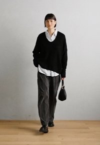 Sweater de malha oversized preto com decote em V, sobre uma camisa branca riscada. Calças cinza soltas e sapatos pretos, segurando uma pequena mala preta.