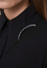 Maglietta nera con un colletto affilato, arricchita da un elemento decorativo metallico di cristalli lungo lo scollo, che aggiunge texture e contrasto.