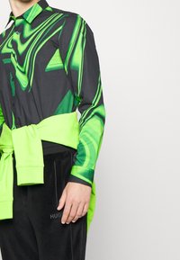 Camicia a motivi neri e verdi con vestibilità slim, dotata di colletto e chiusura a bottoni, abbinata a un maglione verde neon legato in vita.