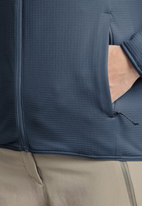 Veste zippée bleu marine en tissu texturé, dotée d'une poche latérale et d'un cordon de serrage réglable à l'ourlet. Associée à un pantalon de couleur claire.