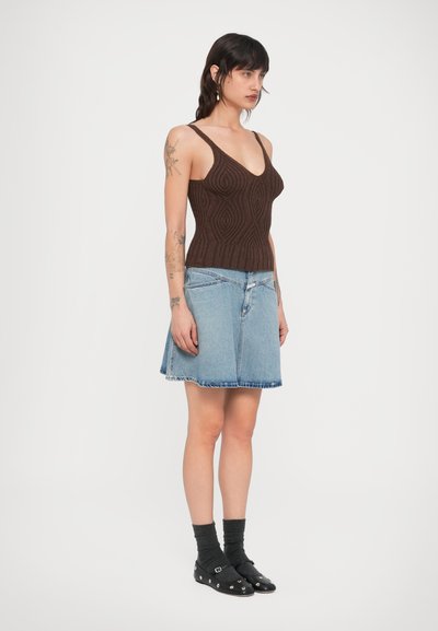 Καφέ πλεκτό τοπ χωρίς μανίκια με V-λαιμόκοψη, συνδυασμένο με φούστα από ανοιχτό μπλε τζιν. Μαύρα επίπεδα παπούτσια με διακοσμητικά μεταλλικά στοιχεία και σκουρόχρωμες κάλτσες.