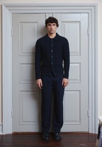 Marineblauwe gebreide cardigan met een kraag en knoopsluiting aan de voorkant, gecombineerd met donkere broeken en zwarte schoenen. Glad textuur, minimalistisch ontwerp.