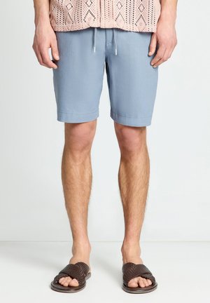 Uomo che indossa pantaloncini azzurri con coulisse, top lavorato a maglia rosa con motivo e sandali intrecciati marrone scuro con dita scoperte, in piedi su pavimento bianco.