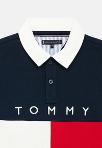 Mornarsko modra polo majica z belim ovratnikom, razporkom z gumbi in napisom "TOMMY" izvezenim z belo barvo na prsih z rdečimi in belimi poudarki.