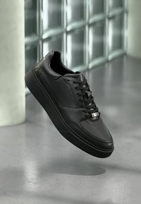 Zwarte leren sneaker met een gladde afwerking, afgeronde neus, geperforeerde accenten, platte veters en een metalen logo aan de zijkant.