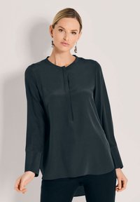 Dunkelgrüne Bluse mit langen Ärmeln, rundem Ausschnitt und einem kleinen Faltenwurf an der Vorderseite. Glattes Material mit einem leichten Glanz, das einen lockeren Schnitt hat.