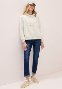 Giovane donna che indossa una felpa grigia chiara, jeans blu arrotolati, sneakers beige e un cappellino azzurro in piedi contro un muro chiaro.