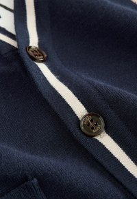 Marine cardigan met witte streep, zachte textuur en donkerbruine knop met de gravure "BAKER". Close-upweergave benadrukt de details.