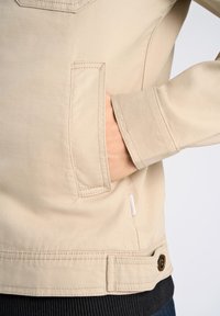 Giacca beige con un design strutturato, dotata di tasca laterale, orlo a coste e dettaglio con bottone in vita. Il materiale appare liscio e resistente.