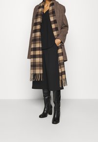 Mantella lunga a scacchi marrone indossata sopra un top e una gonna neri, abbinata a stivali neri con tacco alto e a una sciarpa a scacchi marrone con frange.