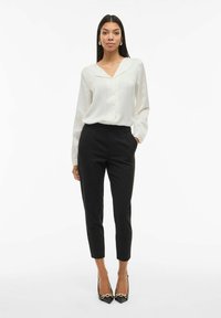 Blusa di seta bianca con scollo a V e maniche lunghe, abbinata a pantaloni neri su misura e tacchi a punta neri con dettagli in catena.