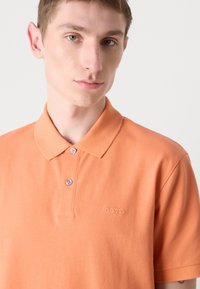 Polo majica kratkih rukava od mekog narančastog pamuka, s klasičnom ovratnikom, dva dugmeta i suptilno izvezenim logom na prsima.