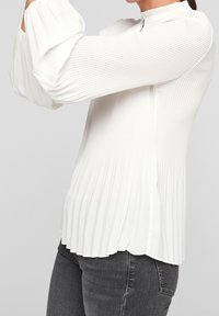 Vit plisserad blus med hög halsringning, puffärmar och dragkedjestängning. Tyget är slätt med subtila vertikala ränder.