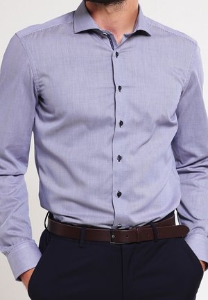 Mężczyzna noszący jasnofioletową koszulę zapinaną na guziki, wpuszczoną w ciemne spodnie z brązowym paskiem, ręce w kieszeniach.