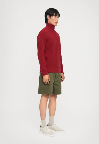 Punane kudumiskootet turtleneck-sweater pooltõmblukuga, millele on lisatud rohelised lühikesed püksid, beežid sokid ja heledad tossud. Mudel seisab külg ees neutraalses taustas.