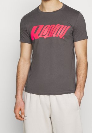 Mies, jolla on tummaharmaa lyhythihainen t-paita, jossa on rohkea punainen tyylitelty teksti, ja vaaleat housut, toinen käsi taskussa.