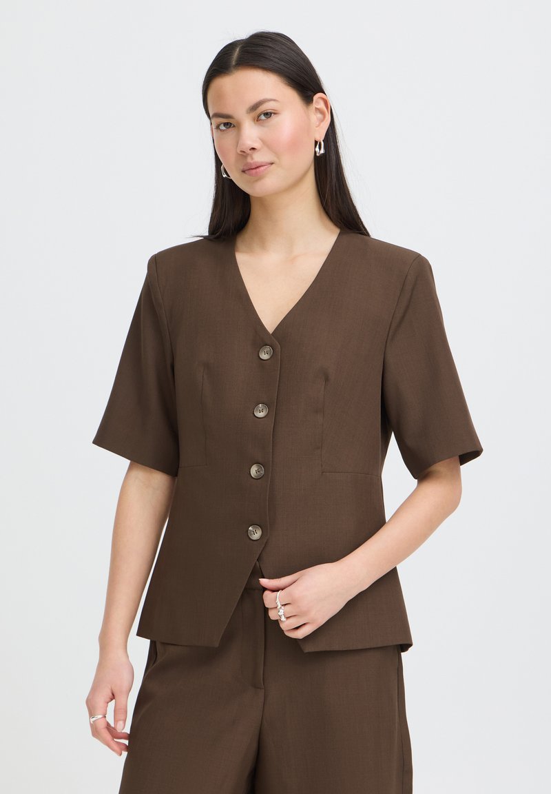 Femme aux longs cheveux foncés portant un blazer marron à manches courtes avec boutons et un pantalon assorti, posant avec la main près de la taille sur fond blanc.