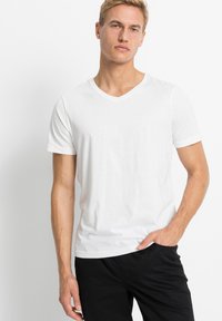 T-shirt bianco a collo a V realizzato in morbido cotone, maniche corte e design aderente, abbinato a pantaloni neri. Texture liscia, senza motivi o decorazioni.