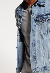 Veste en denim bleu clair avec deux poches poitrine, fermeture à boutons et coutures apparentes, portée par-dessus un polo gris.
