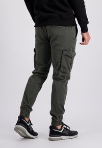 Person som bär olivgröna cargobyxor och svarta sneakers med vit sula, står mot en ljusgrå bakgrund.