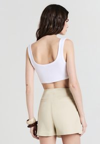 Donna con capelli scuri che indossa un top corto bianco, pantaloni corti beige a vita alta e due braccialetti sul polso sinistro, voltata di spalle.