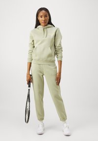 Conjunto de sweatshirt e calças de fato em verde claro, feito de um tecido suave. Apresenta um bolso canguru, punhos canelados e detalhe de logótipo, combinado com uma raquete de ténis.