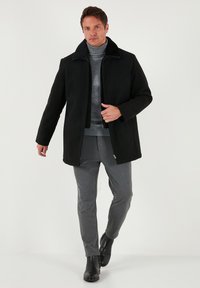 Melnā bez apkakles mētelis ar rāvējslēdzēju, izgatavots no mīksta materiāla. Nēsāts virs pelēka tērpa ar augstu apkakli un pelēkām biksēm, kombinēts ar melniem zābakiem.