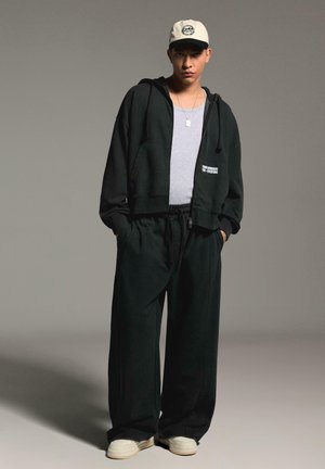 Young man stands with hands in pockets wearing black hoodie, wide black pants, gray shirt, white sneakers, and white baseball cap in studio.