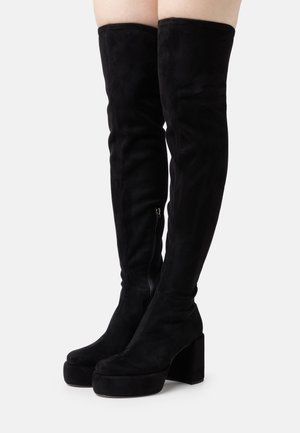 Musta mokkanahkainen korollinen reisiin ulottuva saapas, jossa on paksut kilpikorot ja sivuvetoketjut, käytetty paljaisiin jalkoihin valkoista taustaa vasten.