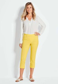 Geel geknipte broek met een gladde textuur en een slank ontwerp, gecombineerd met een witte blouse met lange mouwen. De sandalen hebben een knoopdetail in de band.
