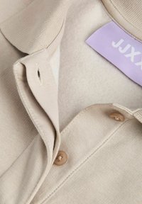 Beige cotton shirt featuring a pointed collar, two buttons, and a lilac label. Smooth texture with a subtle weave pattern.