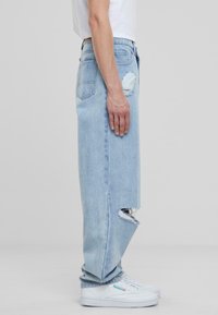Jeans de mezclilla azul claro con corte recto, con un desgarrón en el lado izquierdo y dos bolsillos traseros. Llevados con zapatillas blancas.