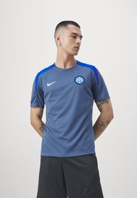 Camiseta gris de manga corta con acentos azules, que presenta un logo circular y el swoosh de Nike en el pecho. Material ligero y transpirable.