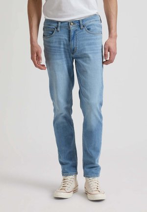 Homme portant un jean slim bleu clair et des baskets blanches, debout devant un fond clair uni.