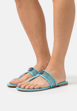 Piedi che indossano sandali piatti turchesi con cinturini trasparenti glitterati e un emblema decorativo a forma di occhio argentato su ciascun cinturino.